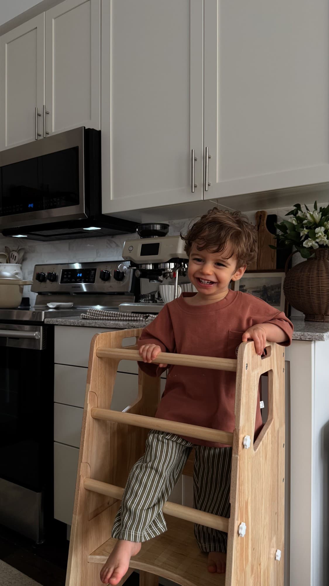 A laughing toddler standing in a natural wood Little Treasures learning tower at the kitchen counter